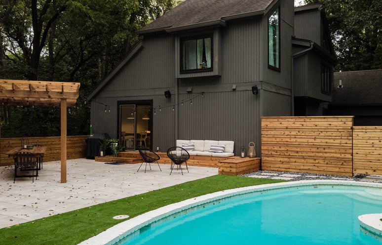 A modern, dark gray house with large windows sits beside a beautifully landscaped backyard featuring a round swimming pool.